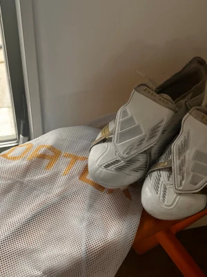 Vita Adidas Predator fotbollsskor - Säljer ett par vita Adidas Predator fotbollsskor med guldiga detaljer och täckt snörning. Skorna har en modern design med tydliga texturer på ovandelen för bättre bollkontroll. Perfekta för dig som vill sticka ut på planen.
