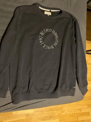 Svart sweatshirt från Blend - Svart sweatshirt från Blend med rund hals och broderad text i cirkel på bröstet där det står 'Blend Originals Since 1993'. Tröjan har en klassisk passform och är perfekt för en avslappnad stil.