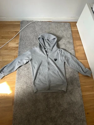 Grå hoodie från Comme des Garçons - Snygg grå hoodie med dragkedja från Comme des Garçons Play. Har huva, två fickor fram och det ikoniska svarta hjärtat med ögon broderat på bröstet. Perfekt för en chill och stilren look. Mjuk bomullskänsla och klassisk passform.