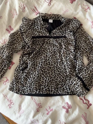 Leopardmönstrad blus med volanger - Snygg blus med leopardmönster och volangdetaljer längs axlar och bröst. Blusen har svarta banddetaljer och är tillverkad i ett lätt, vävt material. Perfekt för dig som vill sticka ut med ett trendigt djurmönster.