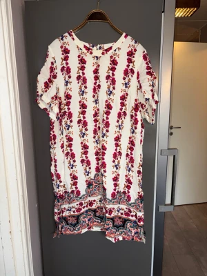 Tunika plussize - Vit tunika  med vertikala ränder av blommor i rosa och blått samt dekorativ kant nertill med paisleymönster. Blusen har rund halsringning och korta ärmar,. 