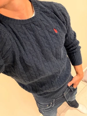 Mörkblå kabelstickad tröja Polo Ralph Lauren - Mörkblå kabelstickad tröja från Polo Ralph Lauren med rund halsringning och röd broderad logga på bröstet. Tröjan har långa ärmar och ribbade muddar vid ärmslut och nederkant. Perfekt för en stilren och klassisk look.