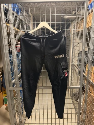 Svarta mjukisbyxor från Hoodrich - Säljer ett par svarta mjukisbyxor från Hoodrich med dragsko i midjan och resår vid bensluten. Byxorna har tryckt logga på benet och en praktisk ficka på sidan med extra detaljer. Perfekta för en avslappnad och streetinspirerad stil.