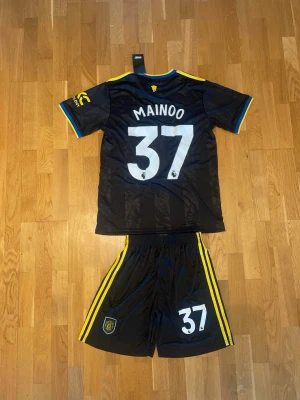 Mainoo Manchester United svart matchställ nr37 - Snyggt svart Manchester United matchställ från adidas med gula detaljer och tryck. Tröjan har namn och nummer (Mainoo 37) på ryggen, klubbmärke och sponsorlogga framtill. Shortsen har gula ränder och klubbmärke. Perfekt för fotbollsträning eller match.