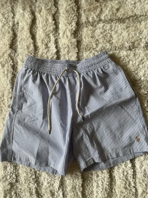Randiga badbyxor från Polo Ralph Lauren - Snygga nästintill oanvända badbyxor med blåvita smala ränder från Polo Ralph Lauren. Klassisk modell med resår och dragsko i midjan samt ficka bak. Liten broderad logga i orange framtill. Perfekta för stranden eller poolen.