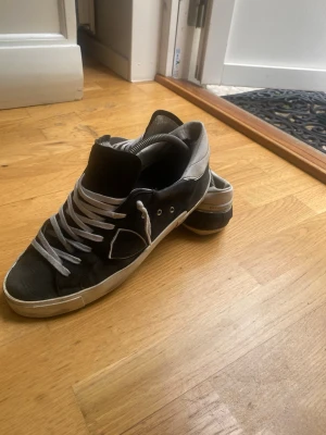 Philipmodel skor  - Snygga svarta sneakers med gråa skosnören och grå häl. Klassisk låg modell med vit sula och diskreta metalldetaljer vid snörningen. Perfekta för en avslappnad streetstil och passar till det mesta i vardagslivet. Ganska slitna längst bak vid sulan. 
