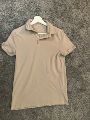 Beige pikétröja från H&M slim fit - Snygg beige pikétröja från H&M i slim fit-modell. Klassisk krage och korta ärmar, perfekt för en clean och stilren look. Tillverkad i mjukt bomullsmaterial som känns skönt mot huden.