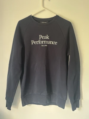 Mörkblå sweatshirt från Peak Performance - Mörkblå sweatshirt från Peak Performance med vit logga och text på bröstet. Tröjan har rund halsringning, långa ärmar och ribbade muddar vid ärmslut och nederkant. Tillverkad i mjukt bomullsmaterial, perfekt för en avslappnad stil.