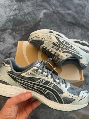Asics Kayano 14 Metropolis/Jasper Green - Snygga sneakers från ASICS i grått och silver med svarta detaljer. Ovandelen är i mesh och syntetmaterial för bra andningsförmåga och komfort. Klassisk låg modell med vit sula och ikoniska ASICS-stripes på sidorna. Perfekta för en sportig och trendig look.