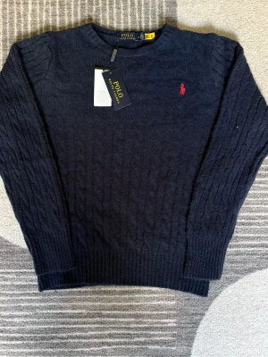 Mörkblå kabelstickad tröja Polo Ralph Lauren - Kabelstickad mörkblå tröja från Polo Ralph Lauren med klassisk röd broderad logga på bröstet. Tröjan har rund halsringning, långa ärmar och ribbstickade muddar. Perfekt för en stilren och tidlös look.
