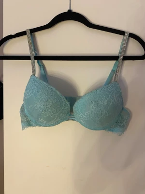 Ljusblå spets-bh med strassband - Snygg ljusblå bh med vadderade kupor och spetsdetaljer. Axelbanden är dekorerade med strass för extra bling. Bh:n har bygel och ger bra stöd. Aldrig använd
