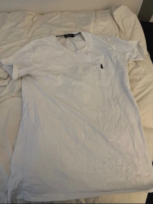 Vit t-shirt från Ralph Lauren - Klassisk vit t-shirt från Ralph Lauren med rund halsringning och korta ärmar. På bröstet finns den ikoniska lilla broderade loggan i svart. Tillverkad i mjuk bomull som känns skön mot huden. Perfekt basplagg till garderoben.