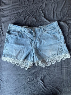 Ljusblå jeansshorts med spetskant - Snygga ljusblå jeansshorts med broderade detaljer och spetskant längst ner. Klassisk femficksmodell med knapp och dragkedja framtill. Perfekta för sommaren och ger en söt touch till din outfit.