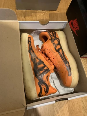 Nike Air Force 1 orange/svart sneakers - Unika Nike Air Force 1 sneakers i orange läder med svart skelettmönster på sidorna. Perfekt för halloween 
