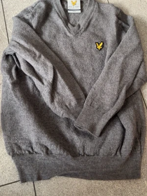 Grå stickad tröja från Lyle & Scott - Grå stickad tröja från Lyle & Scott med V-ringning och klassisk gul logga på bröstet. Tröjan har ribbade muddar vid ärmslut och nederkant. Perfekt för dig som gillar stilren och enkel design.