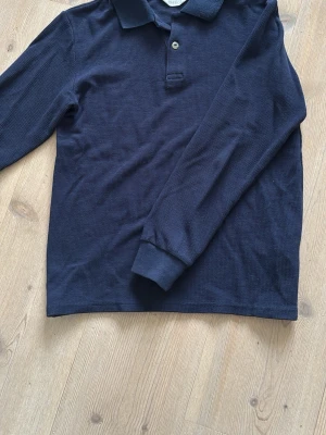 Mörkblå långärmad pikétröja - Snygg mörkblå långärmad pikétröja från H&M med klassisk krage och knappslå framtill. Tröjan är tillverkad i ett strukturerat bomullstyg som ger en cool känsla. Perfekt för en avslappnad men stilren look. Storleken är 152 men passar störte upp till 160