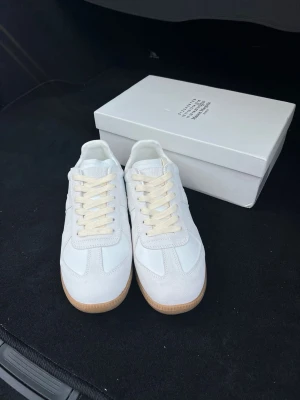 Maison Margiela vita sneakers - Stilrena vita sneakers från Maison Margiela med klassisk låg siluett. Skorna har ovandel i vitt läder och mocka, samt beige snörning och gummisula i naturfärg. Perfekta för dig som gillar minimalistisk och tidlös design.