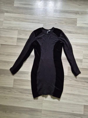 Svart bodycon klänning med lång ärm - Snygg svart bodycon klänning med lång ärm och rund halsringning. Klänningen har insydda paneler i olika svarta material som ger en smickrande siluett. Perfekt för dig som gillar en stilren och modern look.