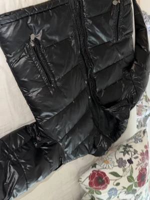 Svart dunjacka från Moncler - Snygg svart dunjacka från Moncler med glansig finish, två dragkedjeförsedda fickor framtill och en ficka med Moncler-logga på ärmen. Jackan har quiltad design och hög krage, perfekt för kalla vinterdagar.