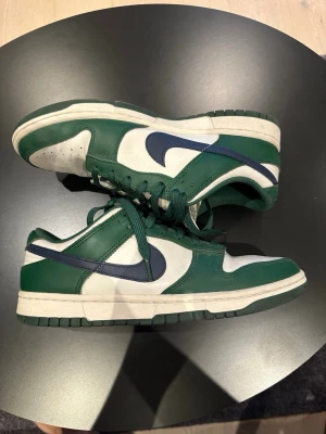 Nike Dunk Low gröna och vita sneakers - Nike Dunk Low sneakers i grönt och vitt med mörkblå swoosh. Ovandel i läder med perforerad tåbox och gröna skosnören. Klassisk siluett med låg profil och vit mellansula. Perfekta för dig som gillar streetwear och sportig stil.