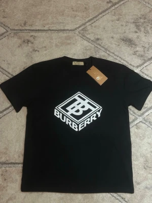 Svart Burberry t-shirt med logga - Svart t-shirt från Burberry med stor vit logga framtill. Klassisk rund halsringning och korta ärmar. Tillverkad i mjuk bomull som känns skön mot huden. Perfekt för dig som vill ha en stilren och trendig look med premiumkänsla.