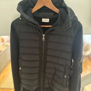 Moncler Cardigan   - Svart pufferjacka/cardigan  från Moncler med stickade ärmar och huva. Jackan har dragkedja framtill, quiltad front och ärmar med Moncler-logga samt ficka med dragkedja. Jackan är självklart äkta, kvitto i pappersform finns kvar samt alla övriga tillbehör som tillkom vid köpet (box, jackfodral osv.). NFC-kod på insidan bevisar ytterligare äktheten. Tveka inte på att hojta till om det finns några frågor eller funderingar!