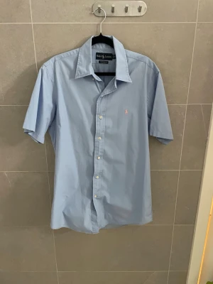 Ljusblå kortärmad skjorta Ralph Lauren - Snygg ljusblå skjorta från Ralph Lauren med korta ärmar och klassisk krage. Skjortan har knappar framtill och en liten broderad logga i rosa på bröstet. Perfekt för en stilren och avslappnad look. skrivas ifall bra bilder krävs. Lite skrinkliga o så men det försvinner i tvätten eller om du ska strika dom eller hur du än gör
