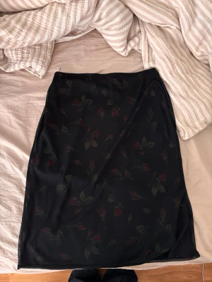 Svart knäkjol med rosor - Svart knäkjol med blommigt mönster av röda rosor och gröna blad. Kjolen har en rak och enkel passform och är tillverkad i ett tunt, lätt material som känns mjukt mot huden. Lappen säger L men känns mer som en M ifall man vill ha den lågmidjat