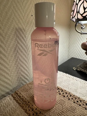 Glittrig bodymist från Reebok - Snygg parfymflaska från Reebok i genomskinligt rosa plast med vit kork. Flaskan har en cylinderformad design och tydlig Reebok-logga tryckt på framsidan. Perfekt för dig som gillar sportiga och fräscha dofter.