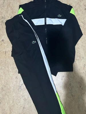 Svart träningsset från Lacoste - Säljer ett svart träningsset från Lacoste med vita och neongröna detaljer. Setet består av en jacka med dragkedja och matchande byxor, båda med Lacoste-loggan. Materialet är lätt och passar perfekt för sport eller chill.