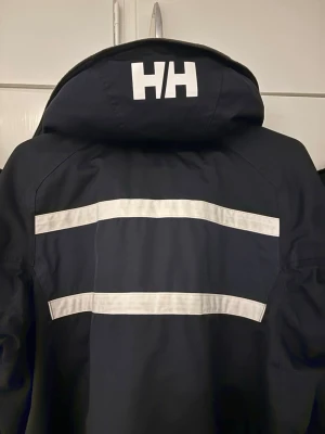vind/seglarjacka från Helly Hansen - Snygg vindjacka från Helly Hansen med vita reflexdetaljer och dragkedja framtill. Jackan har hög krage, praktiska fickor och HH-logga på bröstet. Perfekt för blåsiga dagar och outdoor-aktiviteter. Tveka inte att skriva. Fraktar samma dag.
