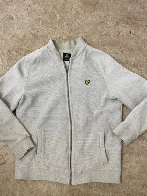 Ljusgrå cardiganjacka från Lyle & Scott - Ljusgrå cardiganjacka med dragkedja från Lyle & Scott. Jackan har ribbade muddar, två fickor framtill och den klassiska gula örnloggan broderad på bröstet. Perfekt för lager-på-lager och enkel att matcha med andra plagg.