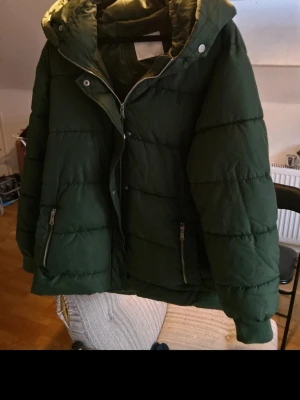 Mörkgrön pufferjacka med huva - Snygg mörkgrön pufferjacka med huva och dragkedja framtill. Jackan har två fickor med dragkedja och quiltad design som ger en cool och modern look. Perfekt för kalla dagar när du vill hålla dig varm och stilsäker. Märket : Samsa collection 