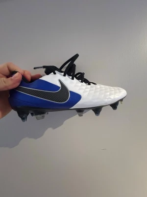 Nike Tiempo Legend 8 Elite SG - Snygga fotbollsskor från Nike i vitt och blått med svart swoosh-logga på sidan. Skorna har en mönstrad ovandel i syntetmaterial och svarta skosnören. Perfekta för dig som vill sticka ut på planen med stilrena och sportiga skor.