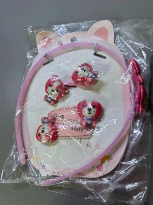 Sött rosa håraccessoarset med hundmotiv - Ett gulligt set med håraccessoarer i rosa plast, inklusive ett diadem och fyra hårspännen, alla dekorerade med små hundar i rosa och vitt med blå overall. Perfekt för att piffa upp frisyren med en lekfull touch.
