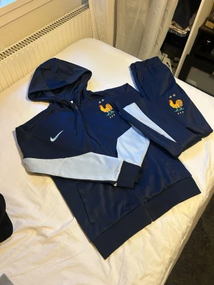 Blå Nike Frankrike träningsset - Snyggt träningsset från Nike med Frankrikes landslagslogga. Setet består av en marinblå huvtröja med dragkedja och matchande byxor, båda med ljusblå detaljer på ärmar och ben. Perfekt för fotbollsträning eller chill. Materialet är polyester.