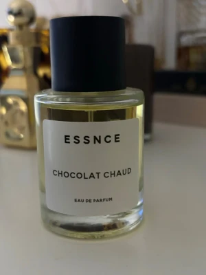 ESSNCE Chocolat Chaud Eau de Parfum - ESSNCE Chocolat Chaud Eau de Parfum kommer i en rund glasflaska med svart lock och enkel vit etikett. Doften har en varm och söt chokladton, perfekt för dig som gillar gourmanddofter. Flaskan är transparent och visar den ljusgula vätskan inuti.