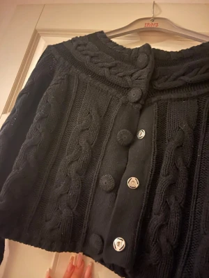 Svart kabelstickad kofta med knappar - Snygg svart kabelstickad kofta med stora dekorativa knappar framtill. Koftan har croppad passform, lång ärm och bred krage med flätmönster. Perfekt för lager-på-lager och en cool vardagslook.