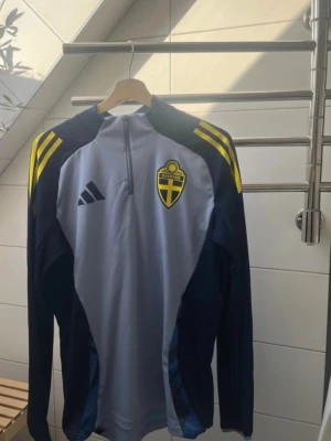 Svenska landslaget träningströja Adidas - Säljer en långärmad träningströja från Adidas med Svenska landslagets emblem på bröstet. Tröjan är grå med mörkblå ärmar och gula detaljer på axlarna. Halv dragkedja framtill och sportig passform, perfekt för fotbollsträning.