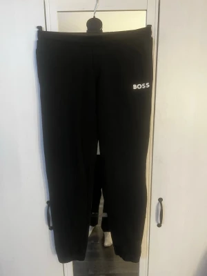 Svarta mjukisbyxor från BOSS - Säljer ett par svarta mjukisbyxor från BOSS med vit logga på vänster lår. Byxorna har en klassisk rak passform och resår i midjan. Perfekta för chill dagar eller när du vill ha en clean och enkel look. Hör av er vid frågor!