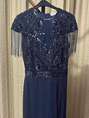 Mörkblå långklänning med fransar - Elegant mörkblå långklänning från lace & beads med glittriga paljetter, blommigt mönster och fransdetaljer över axlarna. Klänningen har rund halsringning och är ärmlös, perfekt för speciella tillfällen.