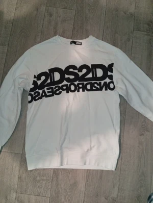 Vit sweatshirt från DS2DS - Snygg vit sweatshirt från DS2DS med stor svart logotyp och text över bröstet. Tröjan har rund halsringning, långa ärmar och ribbade muddar vid ärmslut och nederkant. Perfekt för en avslappnad streetwear-look.