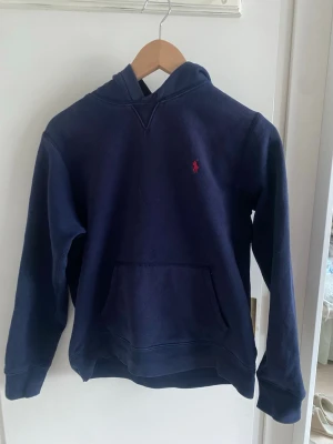 Mörkblå hoodie från Ralph Lauren - Mörkblå hoodie från Ralph Lauren med klassisk huva och känguruficka framtill. Liten röd broderad logga på bröstet. Mjuk bomullskänsla och ribbade muddar vid ärmslut och nederkant. Perfekt för en avslappnad och stilren look.