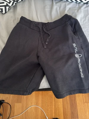 Svarta shorts från Sail Racing - Säljer ett par svarta shorts från Sail Racing med snörning i midjan och tryckt logga på ena benet. Sköna och avslappnade, perfekta för sommaren. Gjorda i mjuk bomull med normal passform.