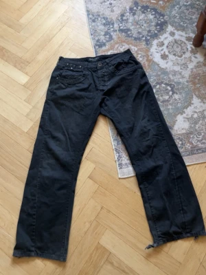 Svarta raka jeans med rå kant - Säljer ett par svarta jeans med rak passform och råa kanter vid bensluten. Jeansen har klassisk femficksdesign och knappgylf. Perfekta för en avslappnad och trendig look.