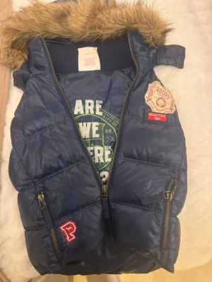 Blå dunväst från PINK XS - Snygg blå dunväst från PINK Victoria's Secret med fuskpäls på huvan och coola patches framtill. Två dragkedjefickor och broderade detaljer ger en sportig vibe. Material: yttertyg i polyester, fyllning av dun och fjäder, foder i bomull och polyester.