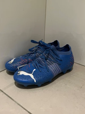 Blåa Puma fotbollsskor med vita detaljer - Säljer ett par blåa fotbollsskor från Puma med vita loggor och silvriga ränder på sidorna. Skorna har snörning, låg profil och små röda detaljer baktill. Ovandelen är i syntetmaterial med struktur för bättre bollkontroll. Perfekta för dig som vill sticka ut på planen.