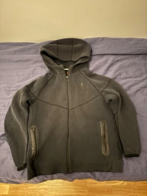 Svart nike tech fleece  - Mysig mörkgrå hoodie i fleece med hel dragkedja framtill och två fickor med dragkedja. Hoodien har en enkel design med markerade sömmar och en stor huva. Perfekt för kalla dagar när du vill vara både bekväm och stilren.