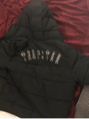 Svart dunjacka från Trapstar - Säljer en svart dunjacka från Trapstar med huva och dragkedja. Jackan har Trapstar-loggan i vitt på ryggen och en rund patch på bröstet. Puffig modell som håller dig varm och har en streetwear vibe. Perfekt för kalla dagar och riktigt snygg till vardagslooken.