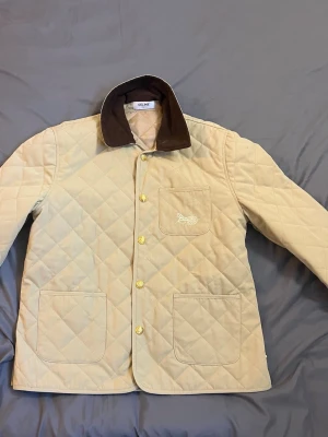 Beige quiltad vindjacka från Celine - Stilren beige quiltad vindjacka från Celine med brunt manchestertyg på kragen. Jackan har tre framfickor, guldfärgade knappar och diskret broderad logga på bröstfickan. Perfekt för dig som gillar klassisk och lyxig stil.